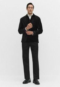 Zwarte corduroy jas met een voorzak, knoopsluiting en een kraag, gecombineerd met zwarte broek en zwarte schoenen. Minimalistisch ontwerp.