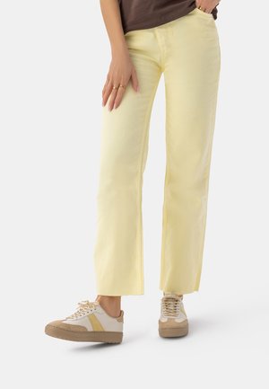 Osoba ubrana w jasne żółte spodnie o prostych nogawkach i beżowe sneakersy z białymi i żółtymi akcentami, stojąca na białym tle.
