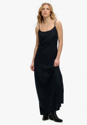 Mujer rubia que lleva un vestido largo negro de tirantes finos y botas negras, sosteniendo el vestido ligeramente levantado con una mano.