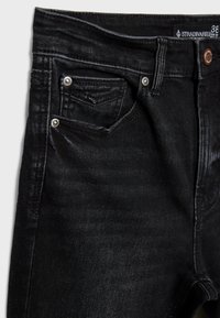 Nahaufnahme von schwarzen Denim-Jeans, die die Vordertasche, Gürtelschlaufe, Metallnieten und einen Knopf mit sichtbarem Markenlabel im inneren Bund zeigen.