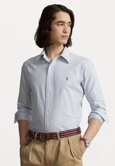 Chemise à rayures bleues et blanches à boutons avec des manches longues, ornée d'un petit logo sur la poitrine et accompagnée d'une ceinture à rayures multicolores.