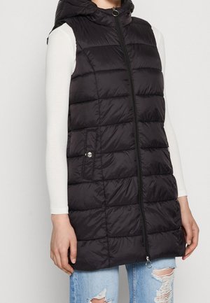 Veste sans manches noire matelassée avec fermeture éclair à l'avant, poches à pression, portée sur un haut blanc à manches longues et un jean bleu clair déchiré.