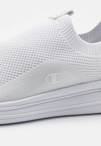 Biały wsuwany sneaker z teksturowaną dzianinową cholewką, gładkim bocznym panelem i białym logo. Posiada miękki kołnierz wokół kostki oraz lekką podeszwę.