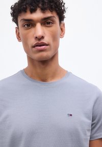 Νεαρός άνδρας με σγουρά μαλλιά που φοράει ανοιχτό γκρι T-shirt με στρογγυλή λαιμόκοψη και μικρό ορθογώνιο λογότυπο στο στήθος, μπροστά σε λευκό φόντο.