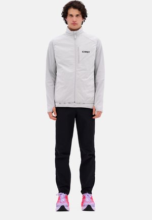 Hellgraue Reißverschlussjacke mit Stehkragen, vorne mit einer Tasche und schwarzem Logo; kombiniert mit schwarzer Hose und mehrfarbigen Sportschuhen.