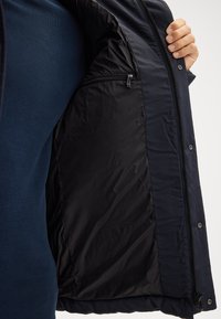 Giacca imbottita navy con zip frontale, che mette in mostra una fodera interna liscia e lucida e una tasca con zip sul lato sinistro.