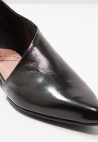 Gros plan sur une chaussure plate à bout pointu en cuir noir brillant, avec une texture visible et une semelle intérieure rose.