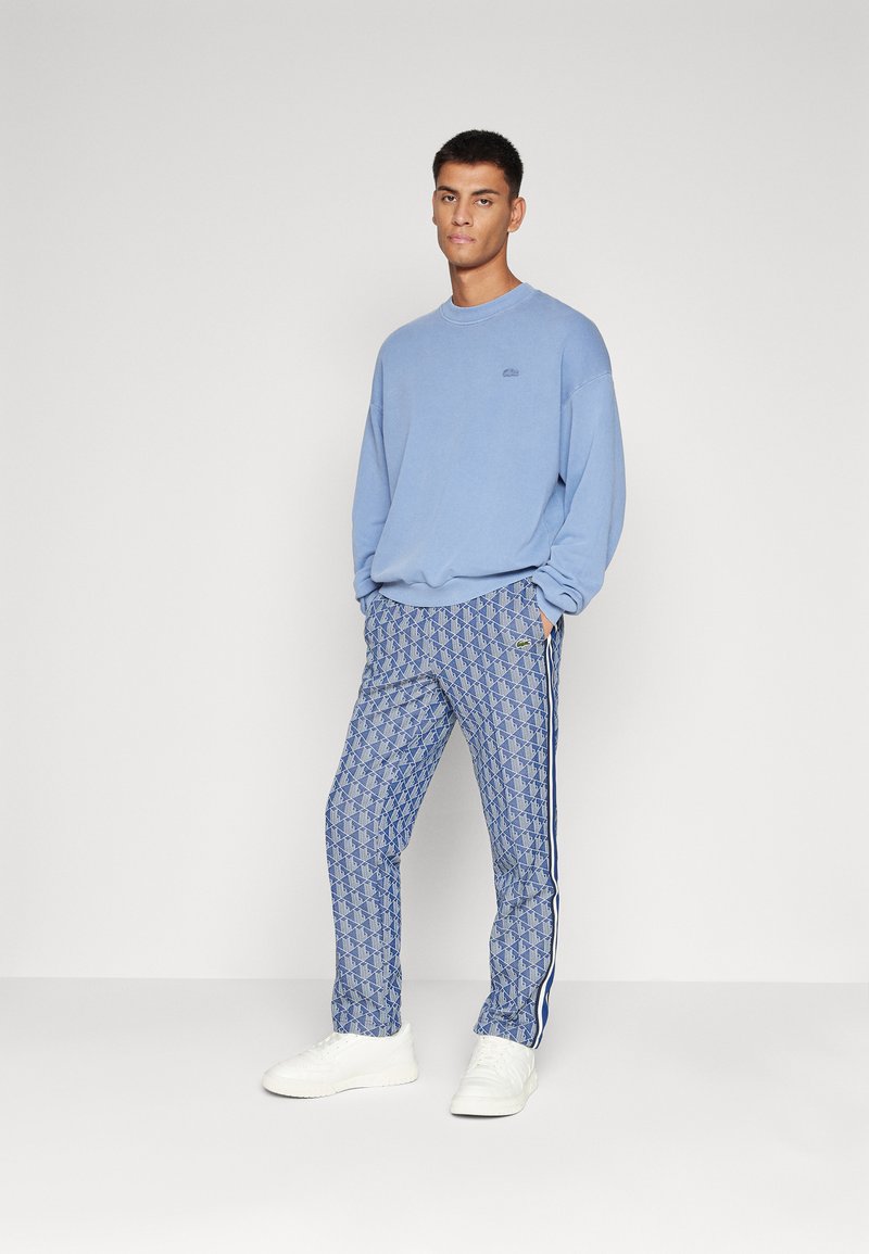 Sudadera azul claro y pantalones estampados en tonos azules con acentos blancos. El modelo está de pie frente a un fondo neutro, vistiendo zapatillas blancas.