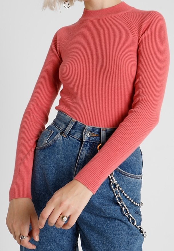 Femme portant un top côtelé corail ajusté à manches longues, rentré dans un jean bleu taille haute avec un accessoire en chaîne argentée.