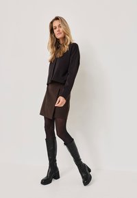 Pull noir court, jupe marron, collants noirs et bottes en cuir noir jusqu'au genou avec un talon épais, posées devant un fond neutre.