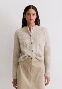 Strick-Cardigan in Creme, mit einem strukturierten Zopfmuster, Rundhalsausschnitt und goldenen Knöpfen. Kombiniert mit einem hellbeigen Rock.