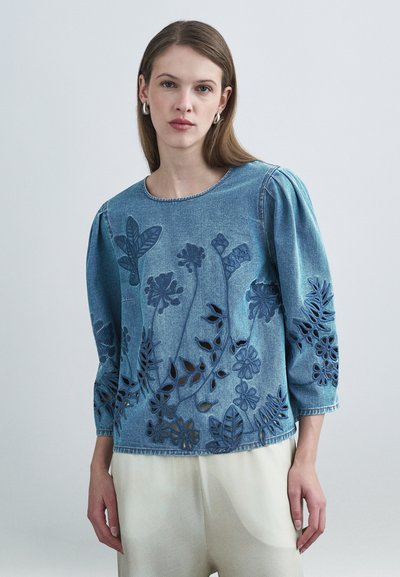 Blouse en denim bleu clair avec motifs floraux découpés, manches trois-quarts et encolure ronde, avec des détails texturés le long des bords.