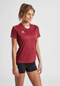 Bordeaux sportshirt met korte mouwen, een V-hals ontwerp en een wit logo. Gecombineerd met zwarte shorts. Glad van structuur, lichte stof.