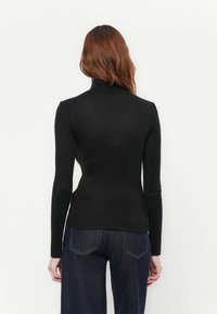 Donna con lunghi capelli rossi che indossa un maglione a collo alto nero aderente e jeans blu scuro, vista di spalle su uno sfondo bianco uniforme.