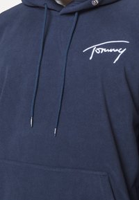 Navyblauwe fleece hoodie met een voorzak, verstelbare trekkoorden en een witte "Tommy" logo op de linkerborst. Zachte textuur.