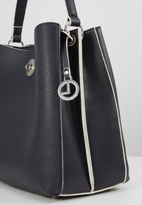 Sac à main en cuir noir texturé avec bordure blanche, fermeture à loquet argentée, pendentif logo rond en argent et poignées noires.