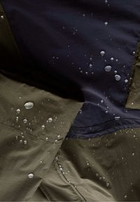 Waterdichte jack met een olijfgroen en marineblauw ontwerp, met zichtbare waterdruppels op de stof, wat de textuur en duurzaamheid benadrukt.