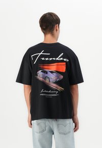 Man wearing black T-shirt with graphic of a purple sports car, orange streak, and "Turbo" text on the back, paired with light blue jeans.