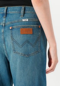 Gros plan sur la poche arrière d'un jean Wrangler bleu avec une étiquette en cuir marron et une main visible reposant sur la hanche.