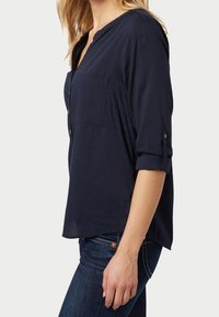 Chemise bleu marine à boutons avec col en V, manches retroussables et poche poitrine, confectionnée en tissu léger à texture subtile.