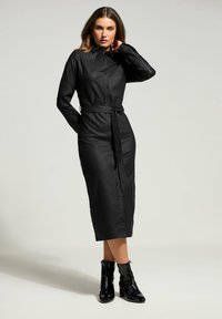 Abito camicia nero a maniche lunghe con colletto, apertura con bottoni e cintura staccabile, realizzato in tessuto liscio, abbinato a stivaletti neri alla caviglia.
