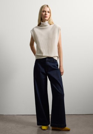 Beige sleeveless turtleneck sweater, paired with wide-leg dark blue denim jeans and bright yellow flats on a neutral background.