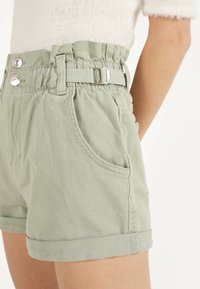 Pantalones cortos de mezclilla verde claro con cintura alta, banda elástica, dos bolsillos delanteros y cinturón en la cintura con herrajes metálicos de acento.