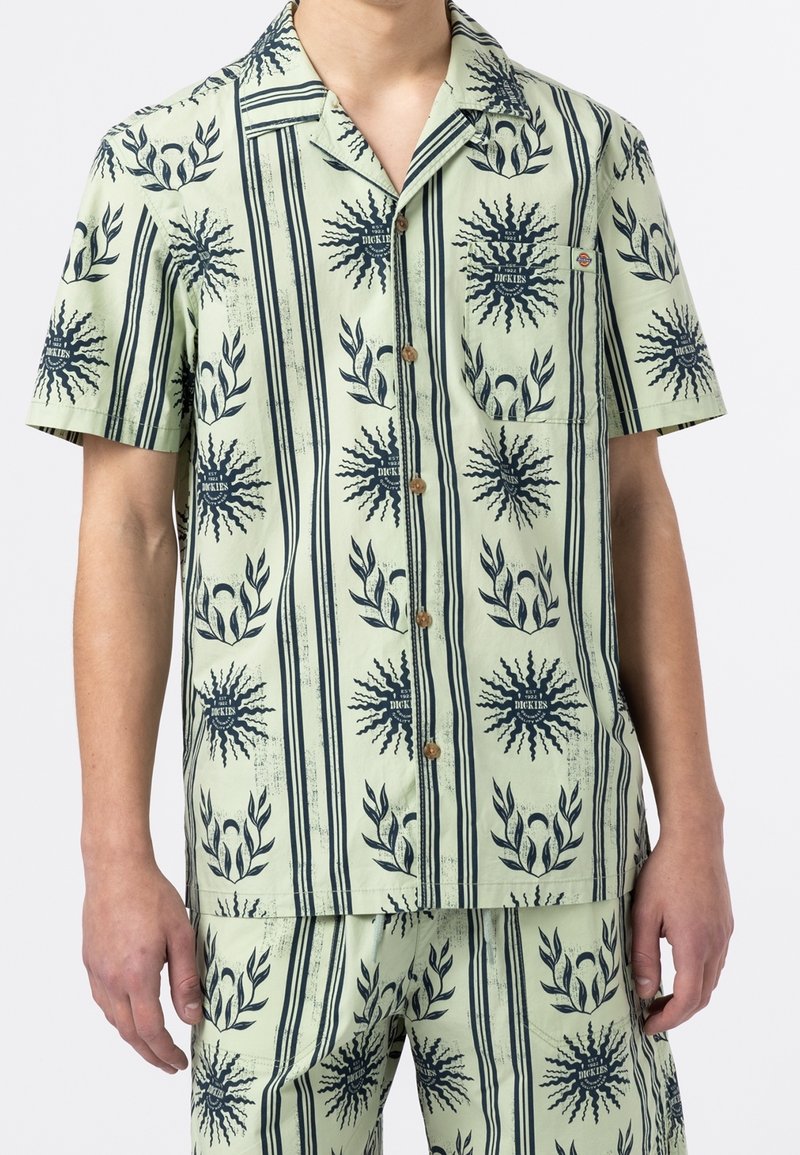 Kortsleeved kragskjorta i ljusgrön tyg med marinblå punktmönster och bladmotiv, med en bröstficka och knappar av trä.