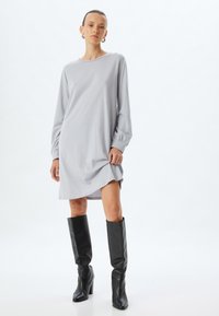Grijze lange mouw jurk met een ontspannen pasvorm, voorzien van een boot neckline en zijsplitten, gecombineerd met kniehoge zwarte leren laarzen.