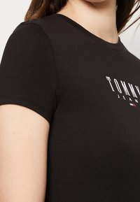 Černé bavlněn�é tričko s krátkým rukávem, přiléhavý design, s bílým logem "TOMMY JEANS" a malým trojbarevným akcentem pod textem.
