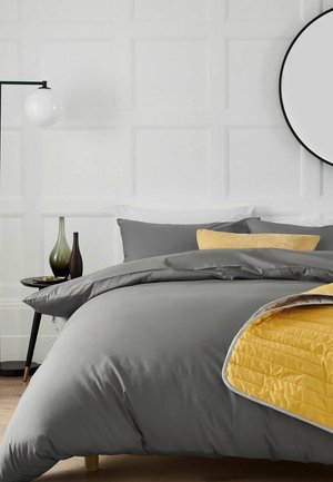 Bett mit grauer Bettwäsche, gelbe Decken-Ecke zurückgefaltet, graue und gelbe Kissen, runder Spiegel, Stehlampe und Beistelltisch mit dekorativen Vasen.