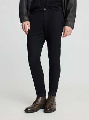 Uomo che indossa pantaloni neri aderenti, stivali in pelle marrone scuro con lacci, camicia nera e giacca in pelle nera, in piedi su sfondo grigio chiaro.
