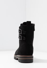 Stivale alla caviglia in suede nero con tre fibbie metalliche, finitura testurizzata, punta arrotondata e suola robusta con un accento marrone chiaro.