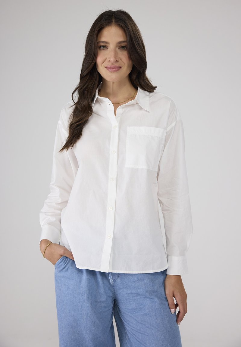 Weißes Hemd mit Knöpfen, kragenferner Ausschnitt, einer Brusttasche und langen Ärmeln. Kombiniert mit hellblauen, weit geschnittenen Hosen.