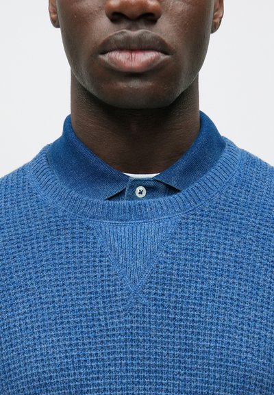 Mėlynas megztinis su tekstūruotu raštu, po juo matosi marškiniai su apykakle. Sagos mėlynos, o apykaklė atitinka megztinio spalvą.