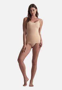Mujer de pie descalza, vistiendo un camisón nude y ropa interior a juego, contra un fondo sencillo.