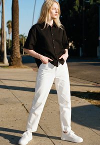 Junge Person mit langen blonden Haaren steht im Freien mit den Händen in den Taschen, trägt ein schwarzes Shirt, weiße Hose und weiße Turnschuhe.