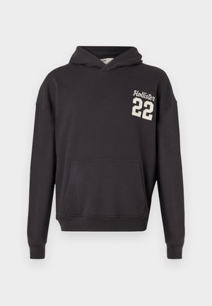 Fekete kapucnis pulóver, elöl zsebbel és krémszínű "Hollister 22" logóval bal mellen, egyszerű fehér háttéren bemutatva.