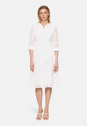 Kvinde iført en hvid midi-kjole med øjelet, 3/4-ærmer, knapper foran og et bundet bælte i taljen, kombineret med beige åben-tå hæle.