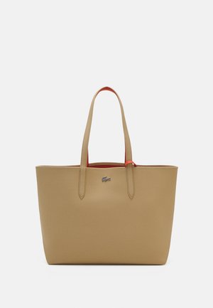 Sac fourre-tout beige avec une texture lisse, deux poignées et un intérieur rouge. Présente un petit logo en argent à l'avant. Design rectangulaire simple.