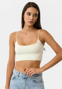 Crème-kleurige cropped tanktop met dunne spaghetti-schouderbandjes en een vierkante halslijn, gemaakt van een gladde, elastische stof. Gekocht in combinatie met lichtblauwe jeans.