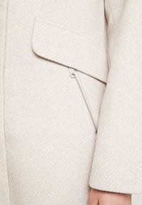 Ljusfärgad, texturerad kappa med en sned framtill ficka och en dragkedjedetalj på sidan. Materialet verkar mjukt med ett fiskbensmönster.