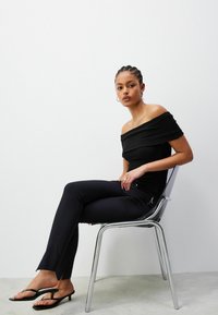 Schwarzes, geripptes Off-Shoulder-Oberteil kombiniert mit engen schwarzen Hosen mit einem seitlichen Reißverschluss, getragen mit schwarzen Flip-Flops auf einem klaren Stuhl.