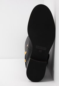 Semelle noire d'une botte en cuir noire avec des détails de boucles dorées, vue par le dessous sur un fond blanc.