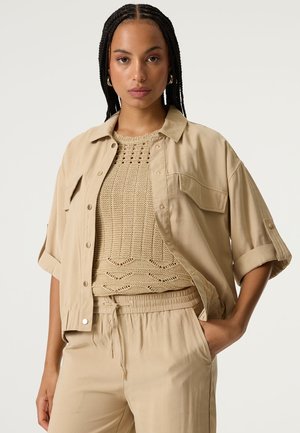 Beiges übergroßes Hemd mit hochgekrempelten Ärmeln, ein gehäkeltes, hellbraunes Oberteil darunter und passende Hosen mit Gummizug an der Taille und seitlichen Taschen.