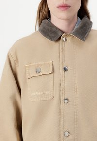 Beige sako s kordurovým límcem, patentkami a kapsou na hrudi, na které je vyšitý nápis "H2OFagerholt" světlou nití.