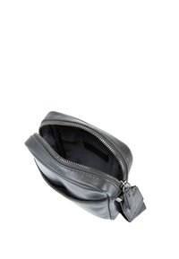 Černá kožená taška se zipovým uzávěrem, má hladkou texturu a připevněnou malou cedulku. Vnitřek je podšit tmavou látkou.