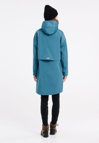 Tyrkysový vodoodolný kabát s kapucňou, s voľným zadným krytom. Skombinovaný s čiernymi legínami a hnedými čižmami. Hladká textúra, minimalistický dizajn.