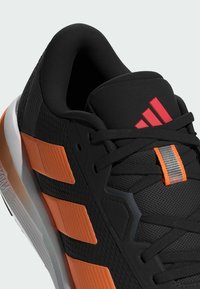 Zwarte sportschoen met gestructureerde mesh bovenwerk, oranje en grijze accenten, gevoerde kraag en kenmerkend rood logo op de tong.