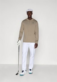 Μπεζ μακρυμάνικο πόλο με λογότυπο, λευκό παντελόνι, λευκά παπούτσια γκολφ με μπλε λεπτομέρειες και λευκό καπέλο γκολφ. Κρατάει ένα γκολφ κλαμπ.